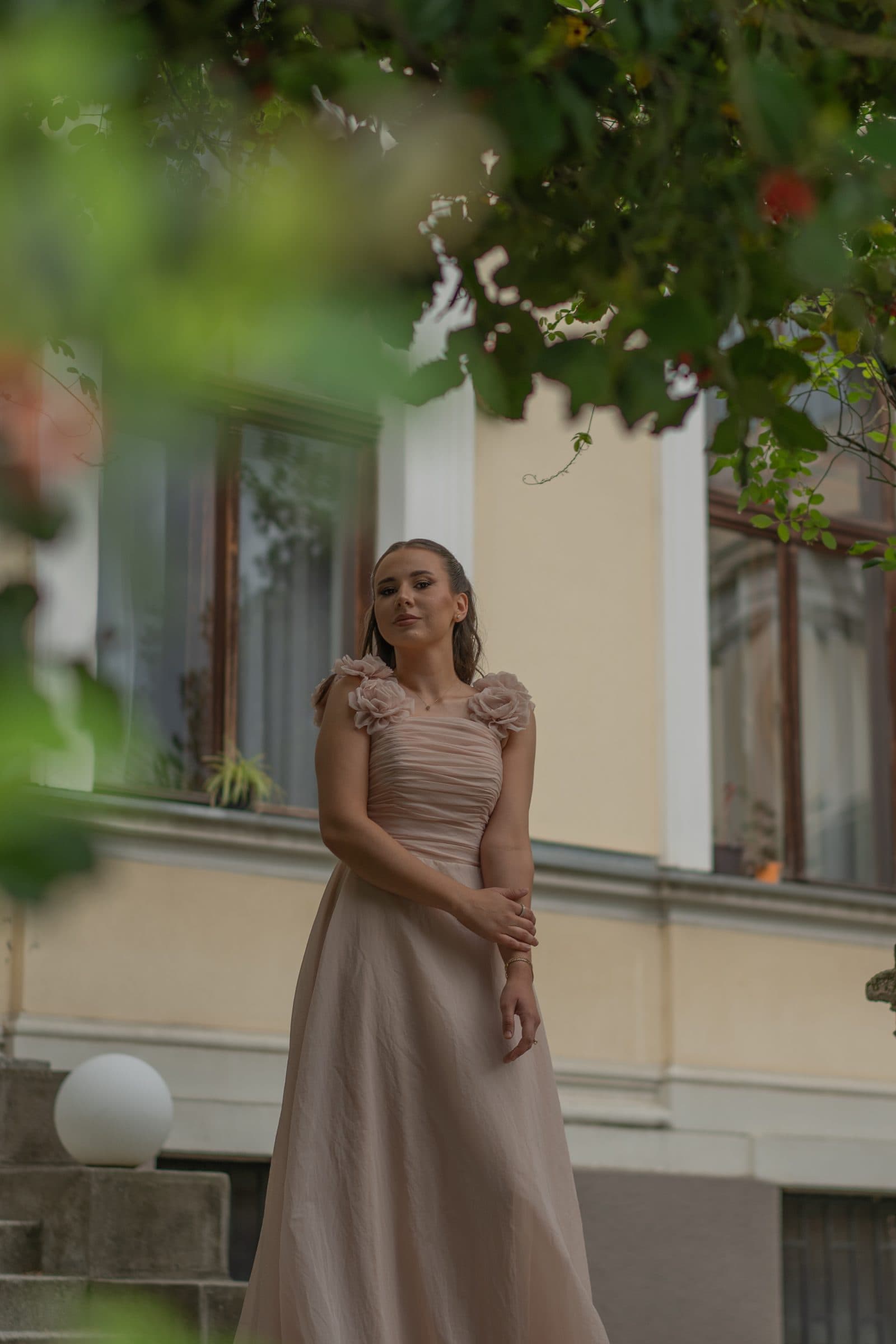 Sumeja in a blush gown under tree branches, looking at the camera